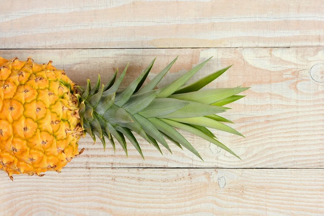 Ananas intero con il ciuffo di foglie verdi appoggiato su un tavolo di legno chiaro