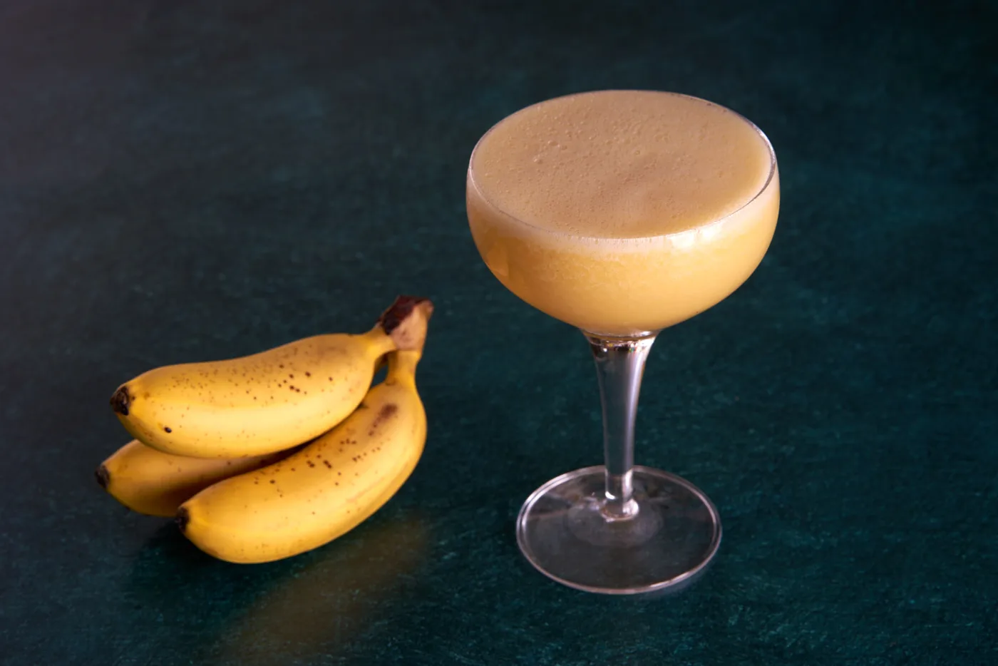Banana Daiquiri cremoso in coppa accanto a un casco di banane su fondo verde scuro