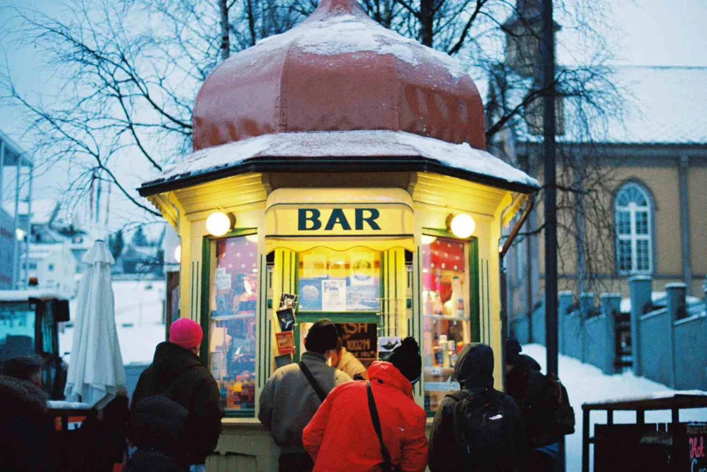 Un chiosco bar con la cupola rossa coperta di neve e l'insegna BAR illuminata al crepuscolo, con persone in giacche invernali in coda davanti