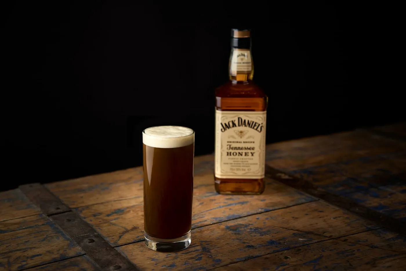 Black Jack: bicchiere alto con drink bruno e schiuma cremosa in stile stout accanto alla bottiglia di Jack Daniel's Honey