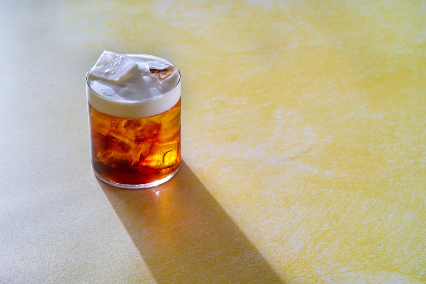 White Russian in tumbler basso con ghiaccio e strato di panna in superficie su fondo giallo