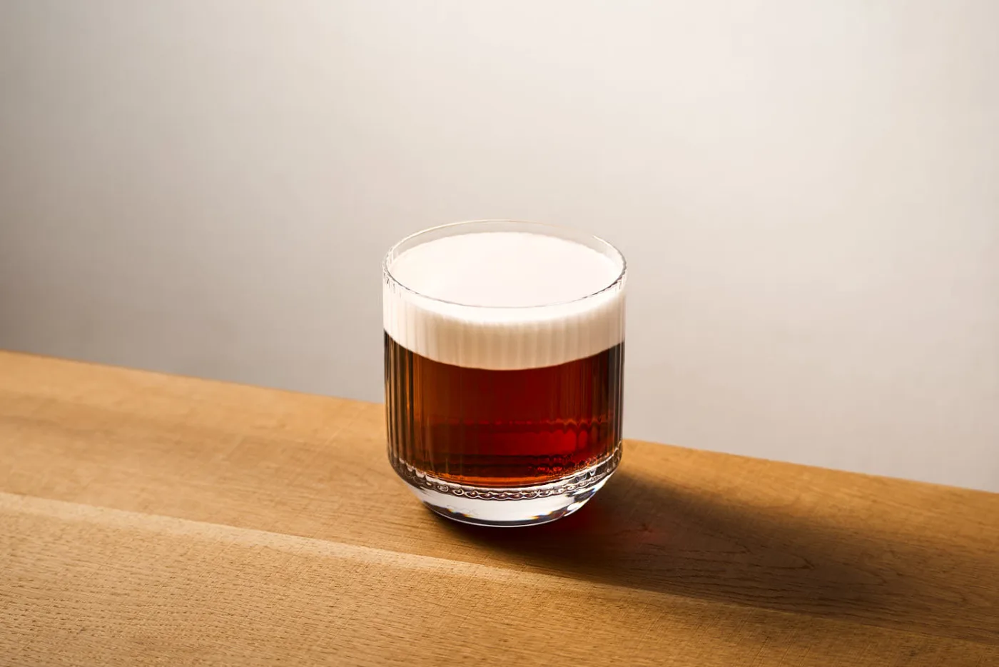 su un tavolo in legno il cocktail white russian preparato con Vodka, liquore al caffè e panna. Il cocktail è servito in un tumbler basso rigato.