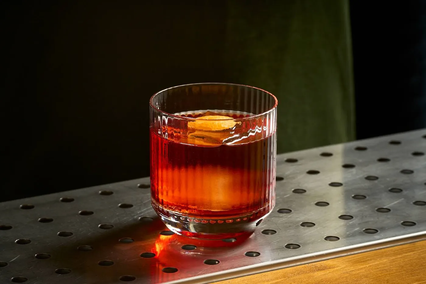 Il Boulevardier servito in un tumbler basso rigato su chunk di ghiaccio, decorato con una scorza di arancia tonda appoggiata sul ghiaccio
