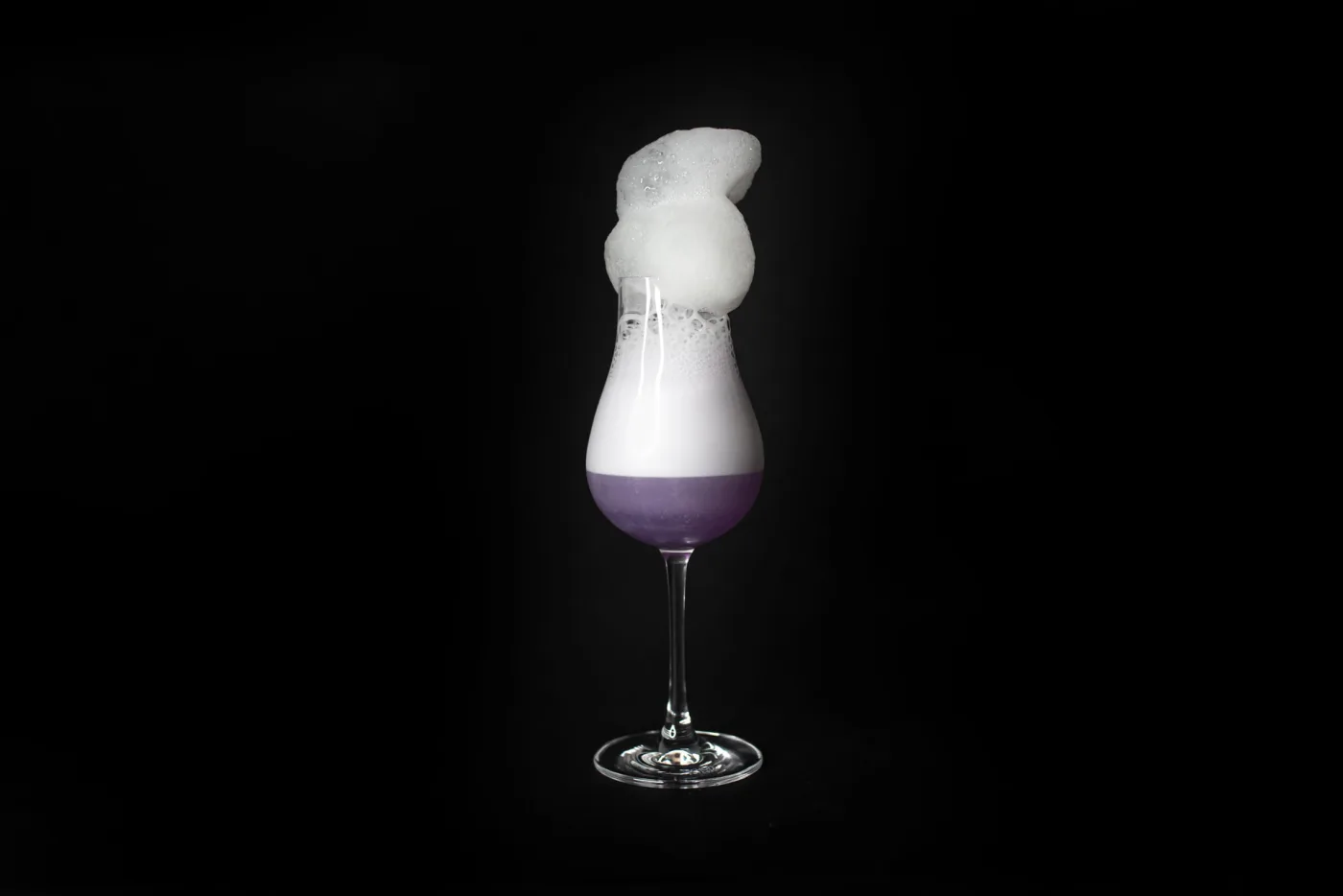 Butterfly Effect: calice da degustazione con drink viola, strato di schiuma bianca e nuvola di mela verde traboccante, su fondo nero