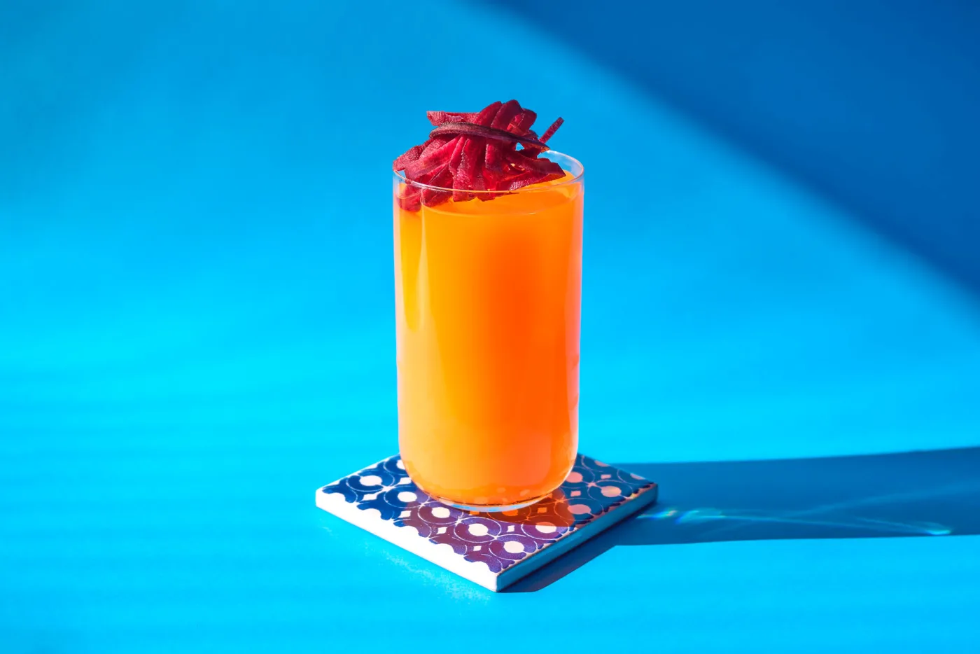 Carrot: long drink arancio vivo in tumbler alto con fili di carota viola, su piastrella decorata e fondo azzurro