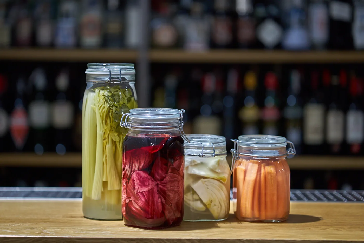 Quattro barattoli con verdure in salamoia in fermentazione lattica — sedano, cavolo rosso, finocchio e carote — sul bancone di un bar