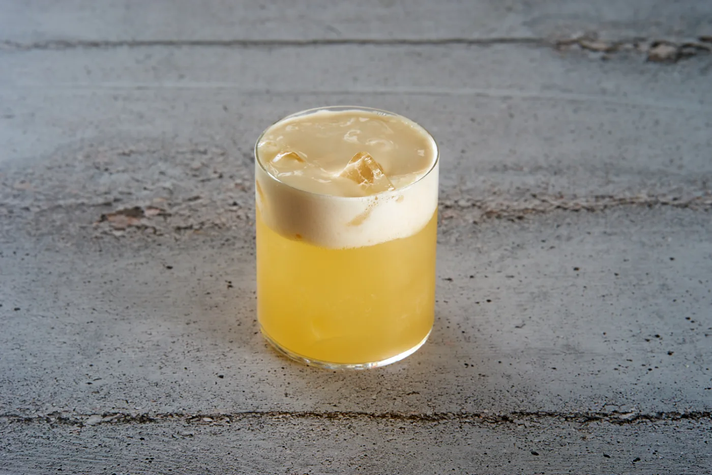 Cherry Sour: tumbler basso con whiskey sour giallo e spessa foam alla ciliegia color crema su piano di cemento