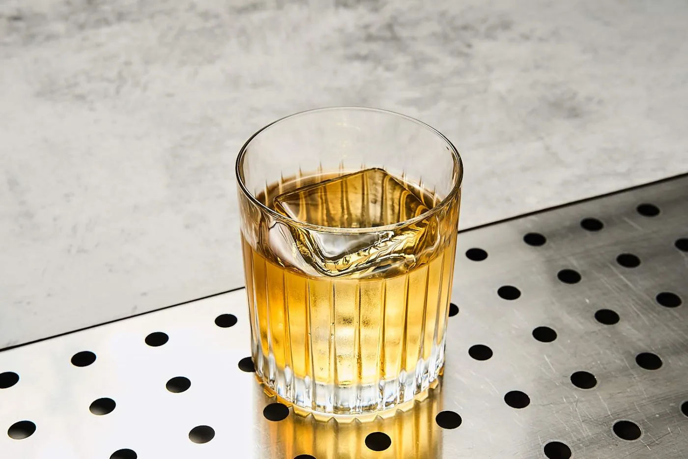 Cocktail ambrato e perfettamente trasparente in un tumbler basso rigato, con un cubo di ghiaccio, su un piano di acciaio forato