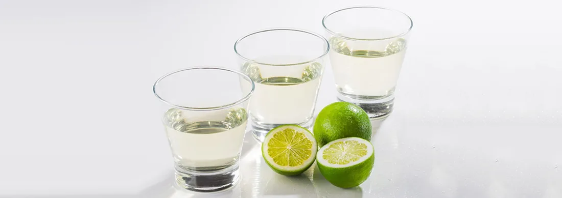 Tre bicchierini di succo di lime chiarificato, perfettamente limpido, accanto a un lime intero e uno tagliato a metà
