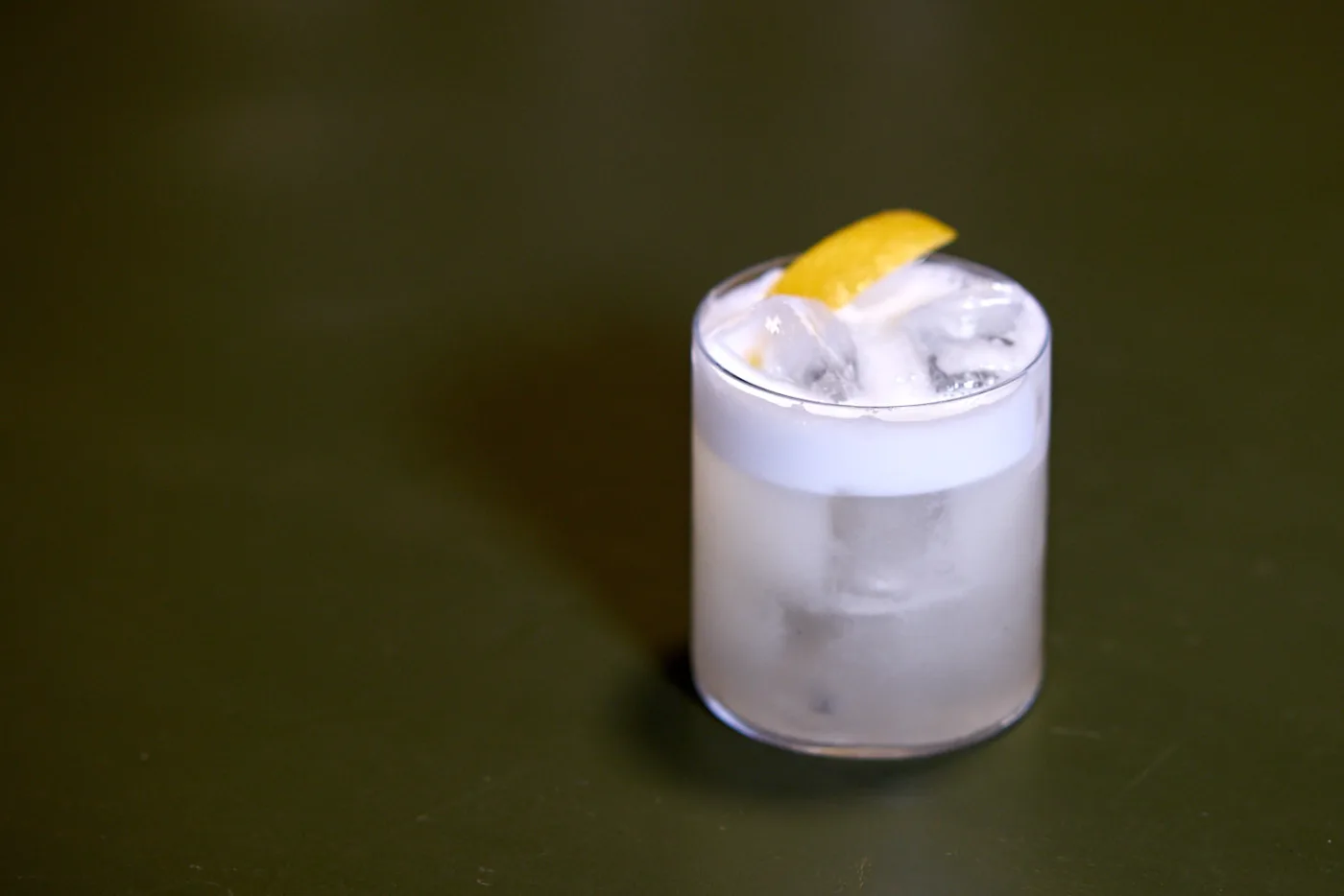 Il Vodka Sour servito in un tumbler basso con ghiaccio, schiuma bianca e scorza di limone, su un piano verde scuro