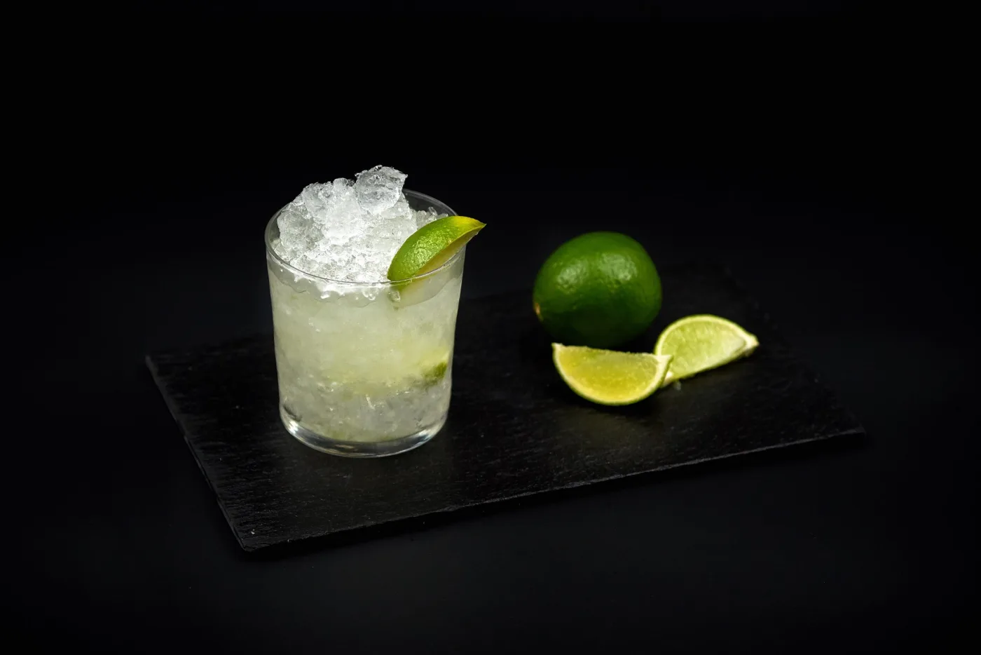 Caipirinha con ghiaccio tritato e spicchio di lime in tumbler basso, su lavagna nera con lime freschi