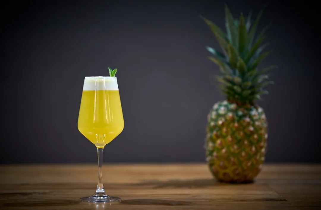 Piña Colada gialla in calice con schiuma bianca compatta e cima di menta, ananas sullo sfondo