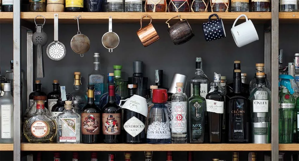Scaffali di legno con decine di bottiglie di gin e distillati allineate e, appesi sopra, colini, tazze di rame e mug da Moscow Mule
