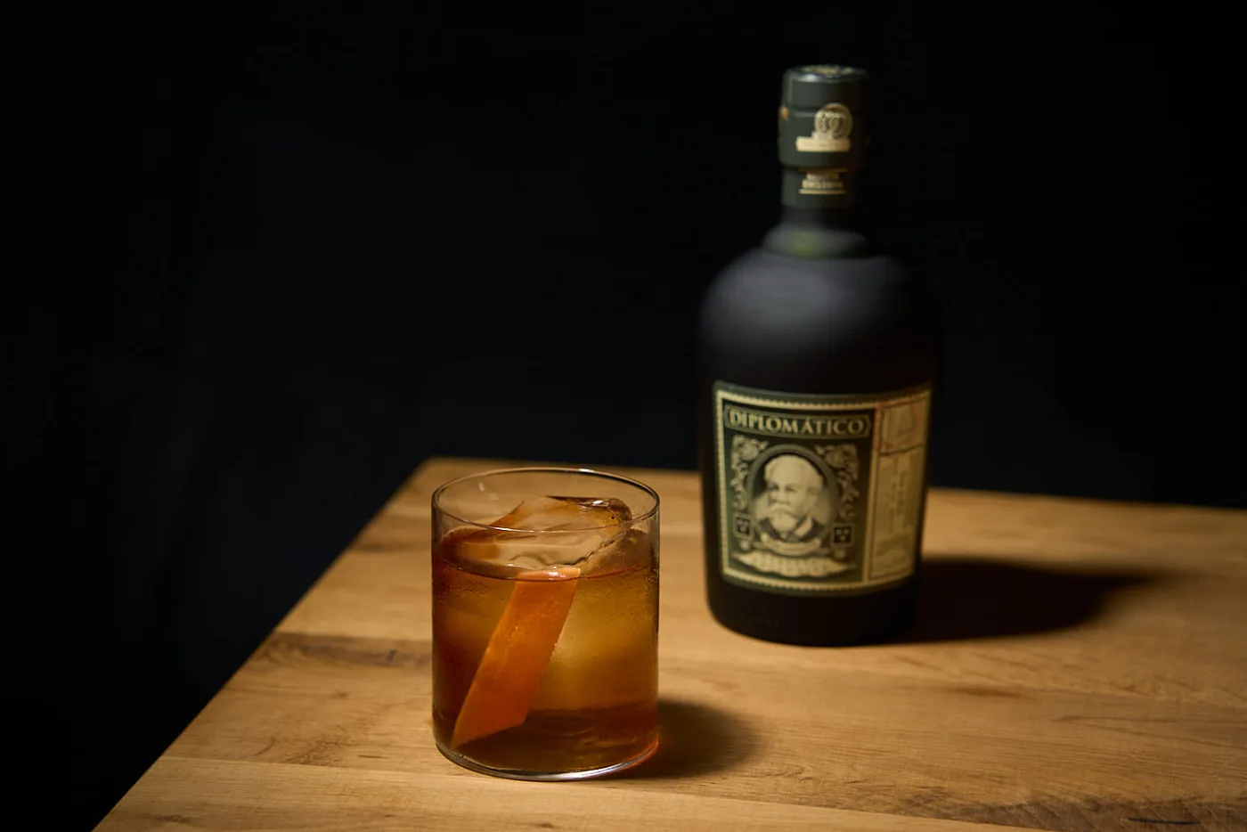 Diplomatico Vecchio Stile: rum old fashioned ambrato su chunk di ghiaccio con twist di arancia accanto alla bottiglia di Diplomatico Reserva Exclusiva