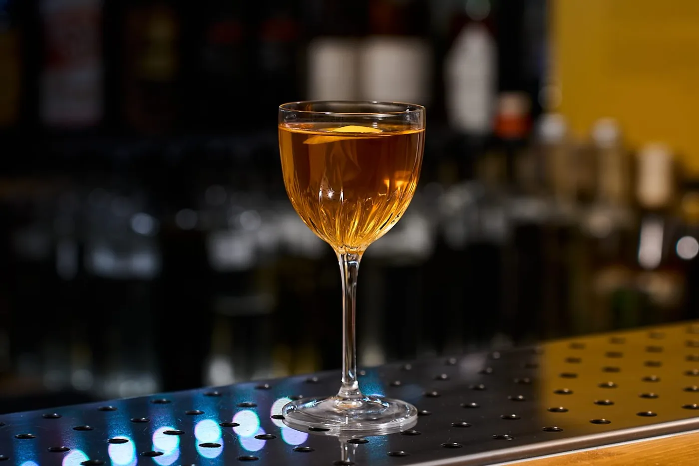 L'East India Cocktail, dal colore ambrato, servito in una coppetta Nick & Nora con motivo in rilievo e scorza di limone, su un banco bar