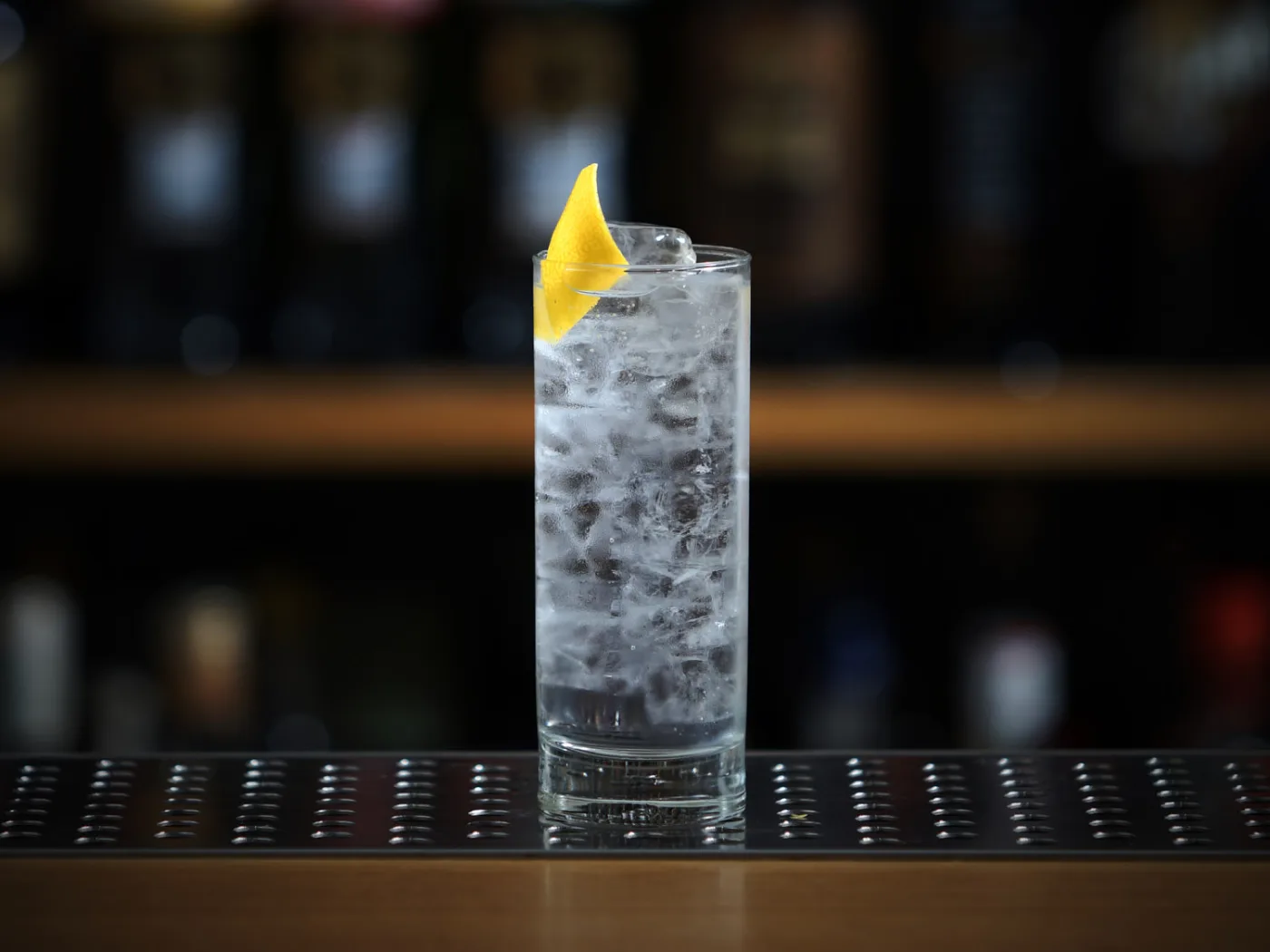 Il Gin Tonic servito in un tumbler alto colmo di ghiaccio con una scorza di limone, su un banco bar