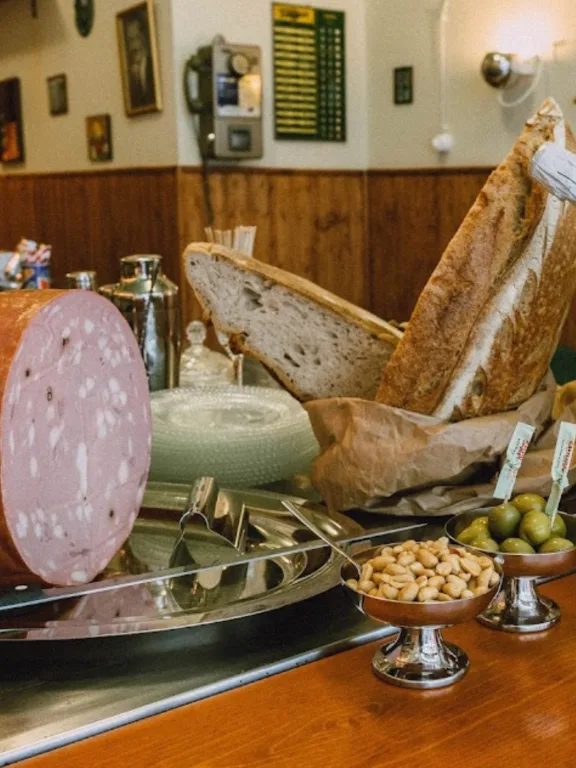 Una parte del bancone del bar Volare di Bologna. Sul banco un vassoio in acciaio con una grossa Mortadella, alcuni snack, due pagnotte e una bowl contenente tre bottiglie di Champagne.