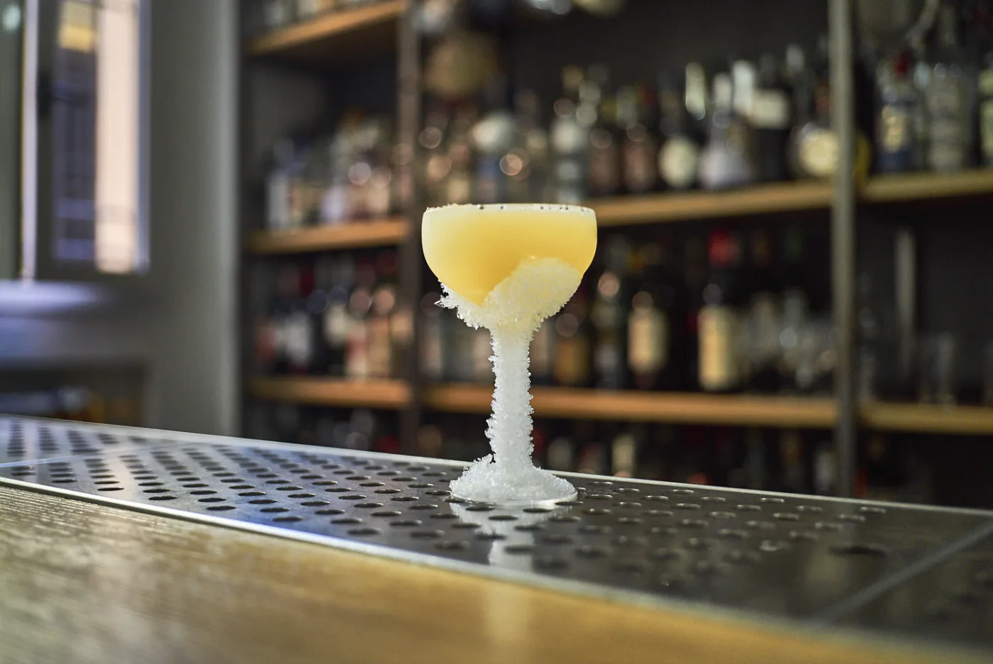 Frisco Sour giallo in coppetta ghiacciata su banco bar con bottiglie sfocate sullo sfondo