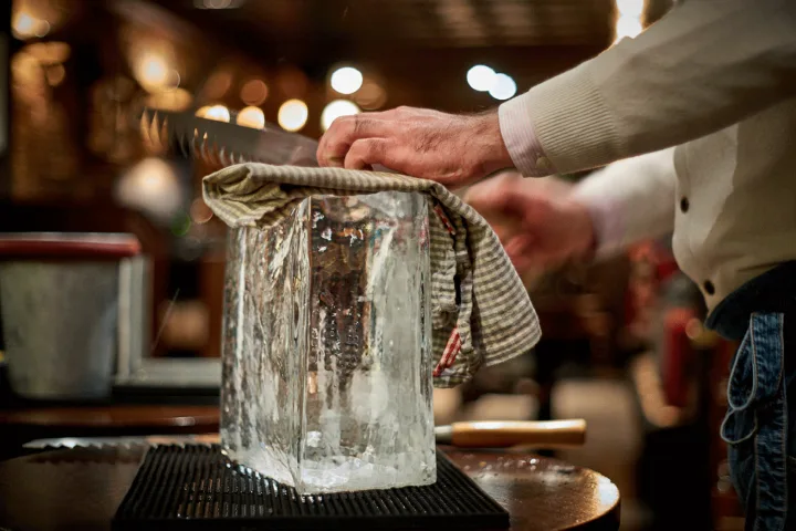 Bartender che lavora un grande blocco di ghiaccio trasparente con una sega, dietro il bancone di un bar