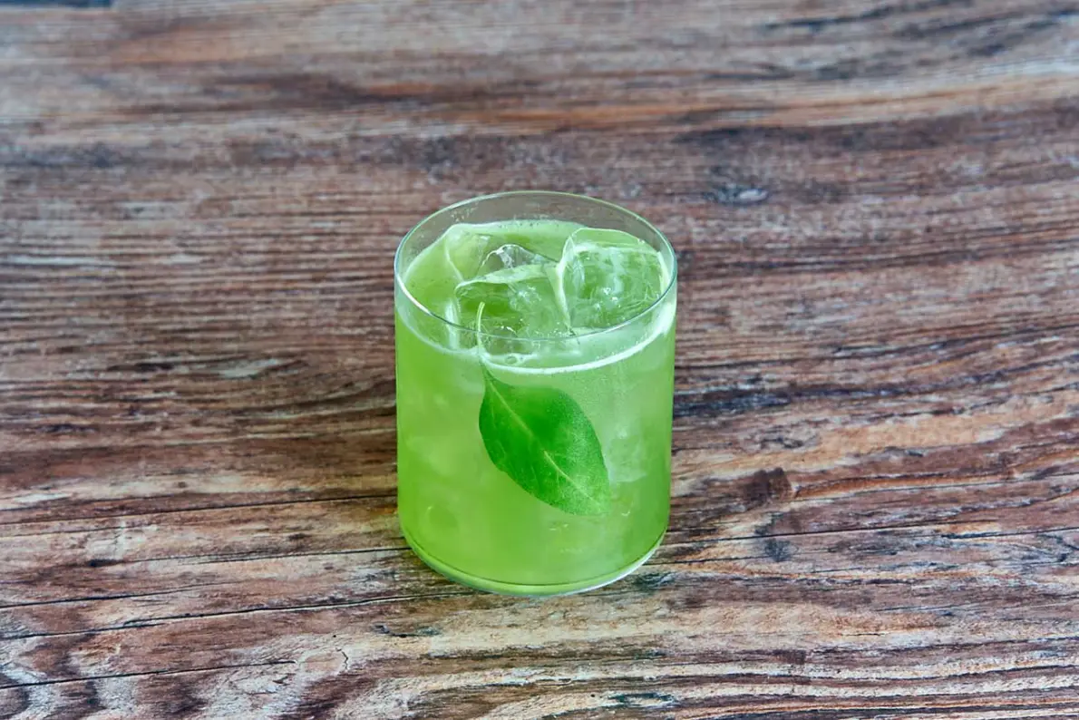 Gin Basil Smash verde brillante in tumbler basso con ghiaccio e foglia di basilico su tavolo di legno