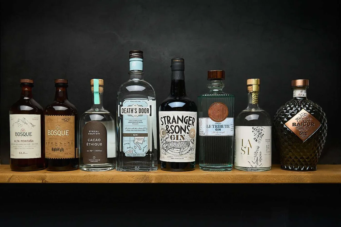 Otto bottiglie di gin di produttori diversi allineate su una mensola di legno, su fondo nero