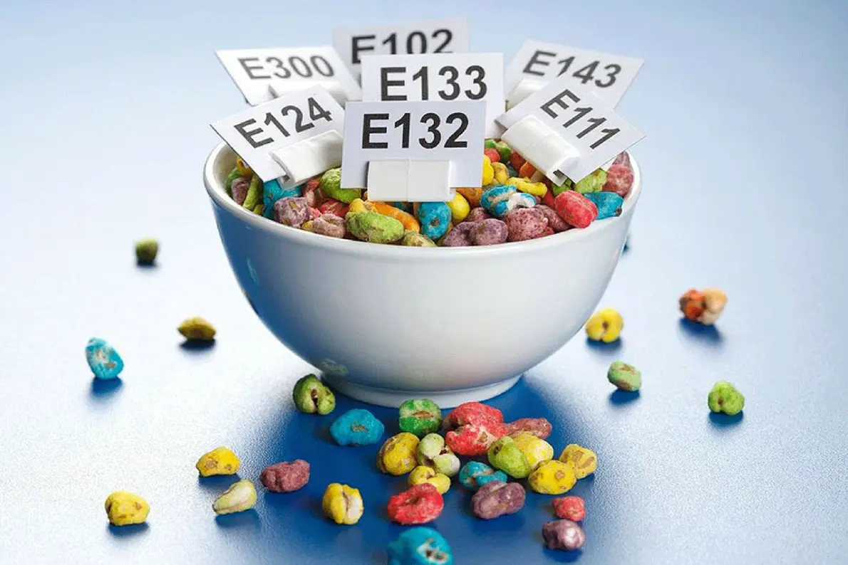 Una ciotola di cereali colorati con dentro dei cartellini con le sigle degli additivi E102, E124, E132, E133, E143 e E300