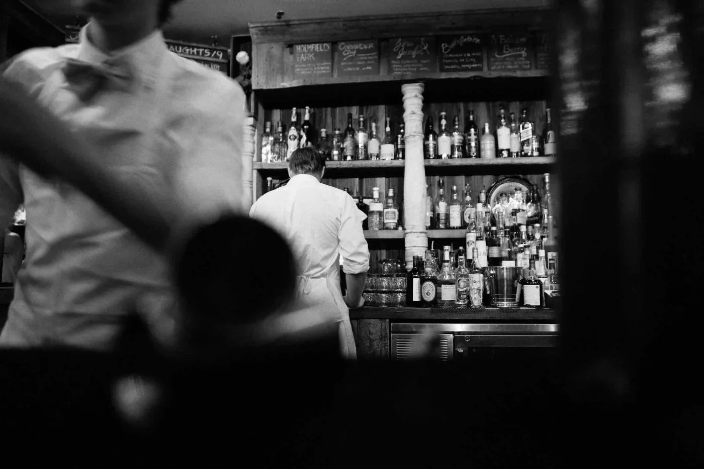 Fotografia in bianco e nero di un bar: in primo piano, sfocato, un barman con camicia bianca e papillon, sullo sfondo un collega di spalle davanti allo scaffale delle bottiglie