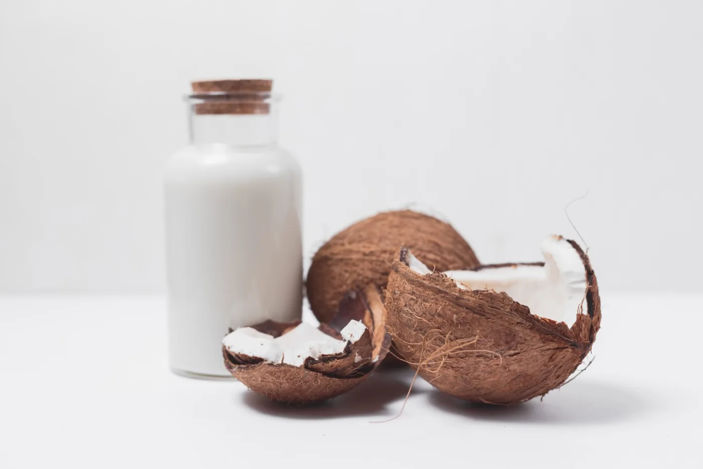 Una bottiglietta di latte di cocco con tappo di sughero accanto a una noce di cocco aperta, su fondo bianco