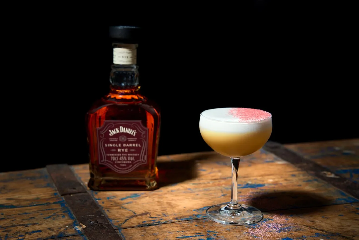 Jack and the Bees: sour giallo in coppetta con schiuma bianca e polvere di fragoline di bosco rosa, accanto alla bottiglia di Jack Daniel's Single Barrel Rye