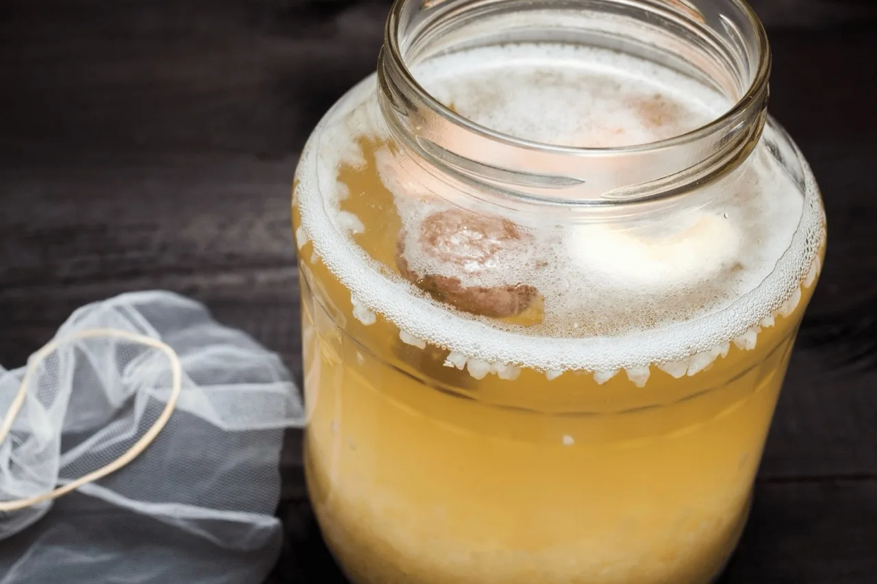 Barattolo di vetro con kefir d'acqua in fermentazione, frutta essiccata a galla e telo traspirante con elastico accanto