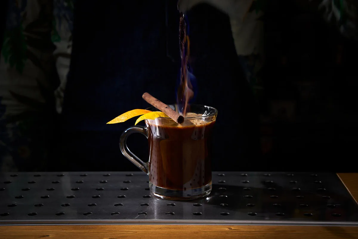 Kona Coffee Grog fiammeggiante in tazza di vetro con stecca di cannella e scorze di agrumi sul bordo
