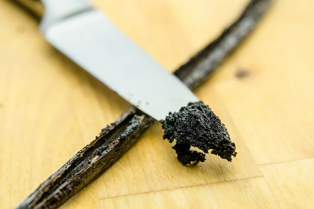 Un baccello di vaniglia aperto su un tagliere di legno, con i semi neri raccolti sulla lama di un coltello
