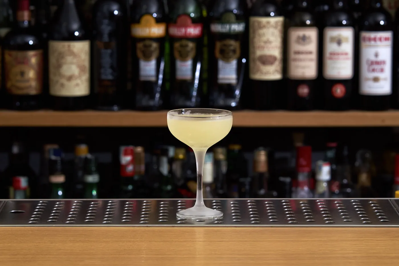 Last Word giallo-verde pallido in coppa brinata su banco bar con bottiglie sfocate