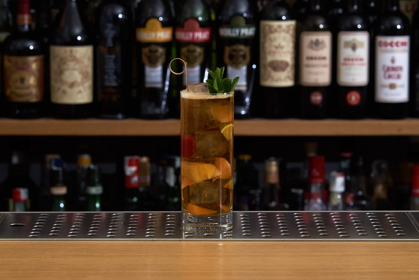 Il Pimm's Cup in un tumbler alto con frutta fresca, ghiaccio, ciuffo di menta e stecchino, davanti alla bottigliera del bar