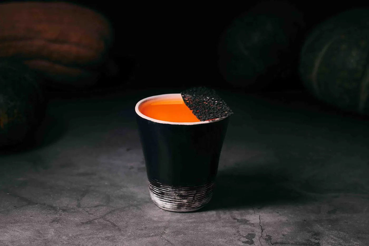 PumpKinG: bicchiere di ceramica nera con drink arancio vivo alla zucca e cialda scura, con zucche sullo sfondo
