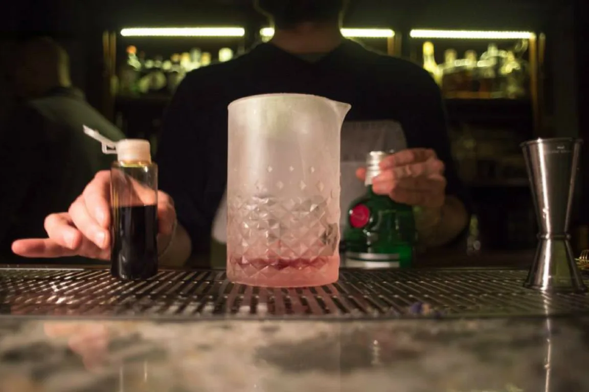 Mixing glass rigato completamente brinato appoggiato sul bancone del bar, con jigger e bottiglie sullo sfondo
