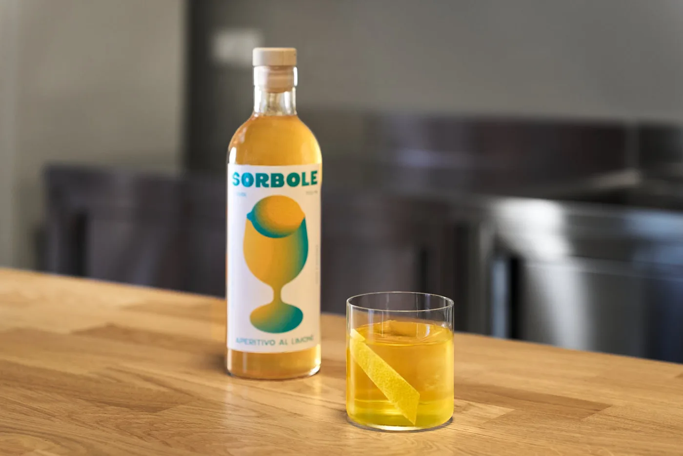 Su un banco bar Il cocktail Negroni Giallo servito in un bicchiere tipo tumbler basso con un chunk di ghiaccio e decorato con una scorza di limone. Accanto al cocktail una bottiglia di Sorbole, aperitivo al limone.