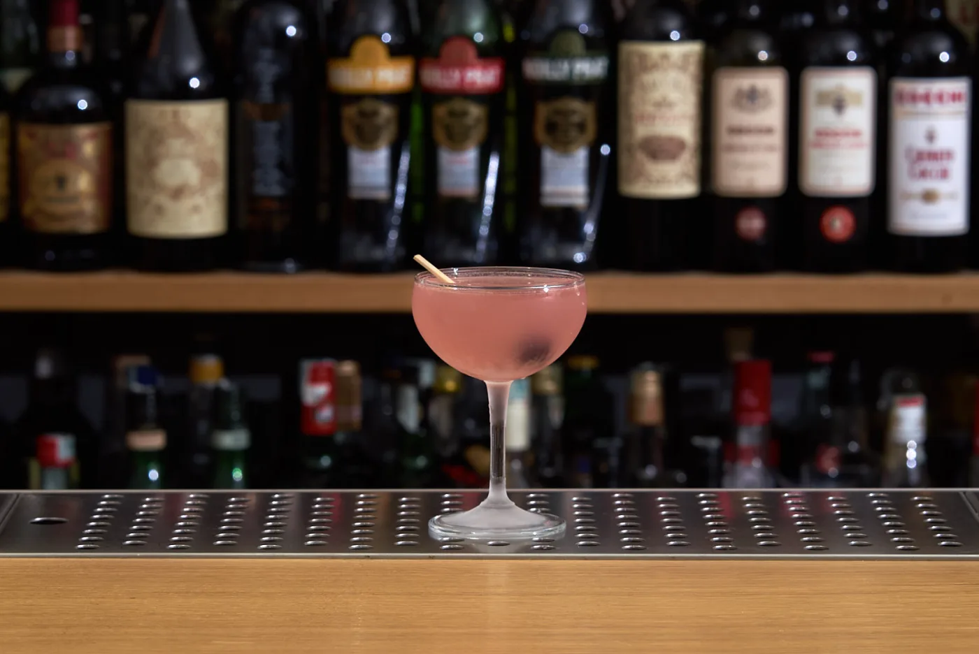 Cosmopolitan rosa in coppa brinata con stecchino, su banco bar con bottiglie sfocate