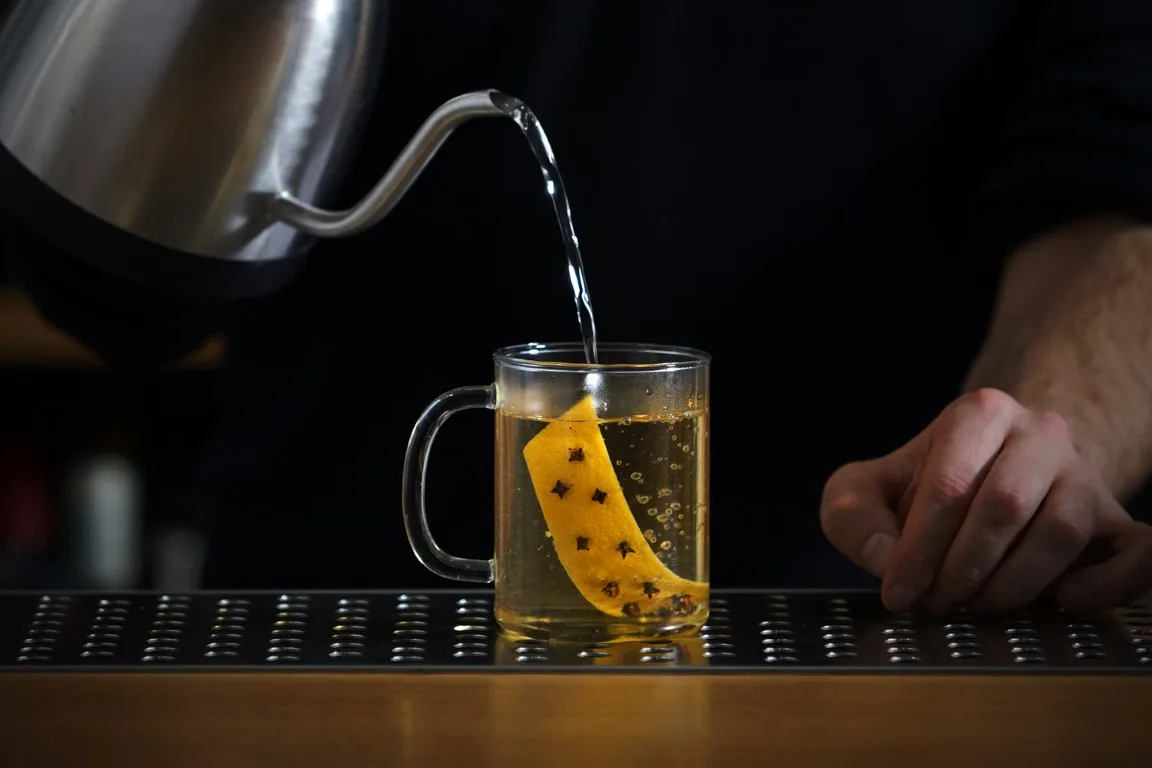 Acqua calda versata dal bollitore in una tazza di vetro con scorza di limone steccata di chiodi di garofano