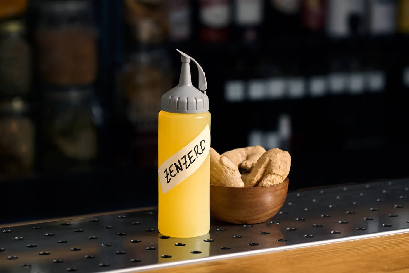 Squeeze bottle gialla con etichetta scritta a mano Zenzero contenente sciroppo di zenzero home made, su un banco bar accanto a una ciotola di legno con radici di zenzero fresco