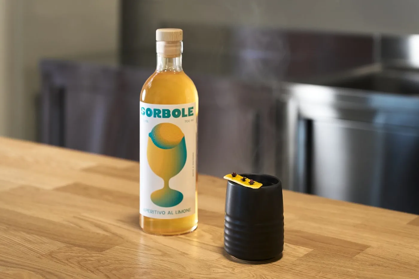 Sorbole Hot Toddy fumante in mug nera con scorza di limone e chiodi di garofano accanto alla bottiglia di Sorbole