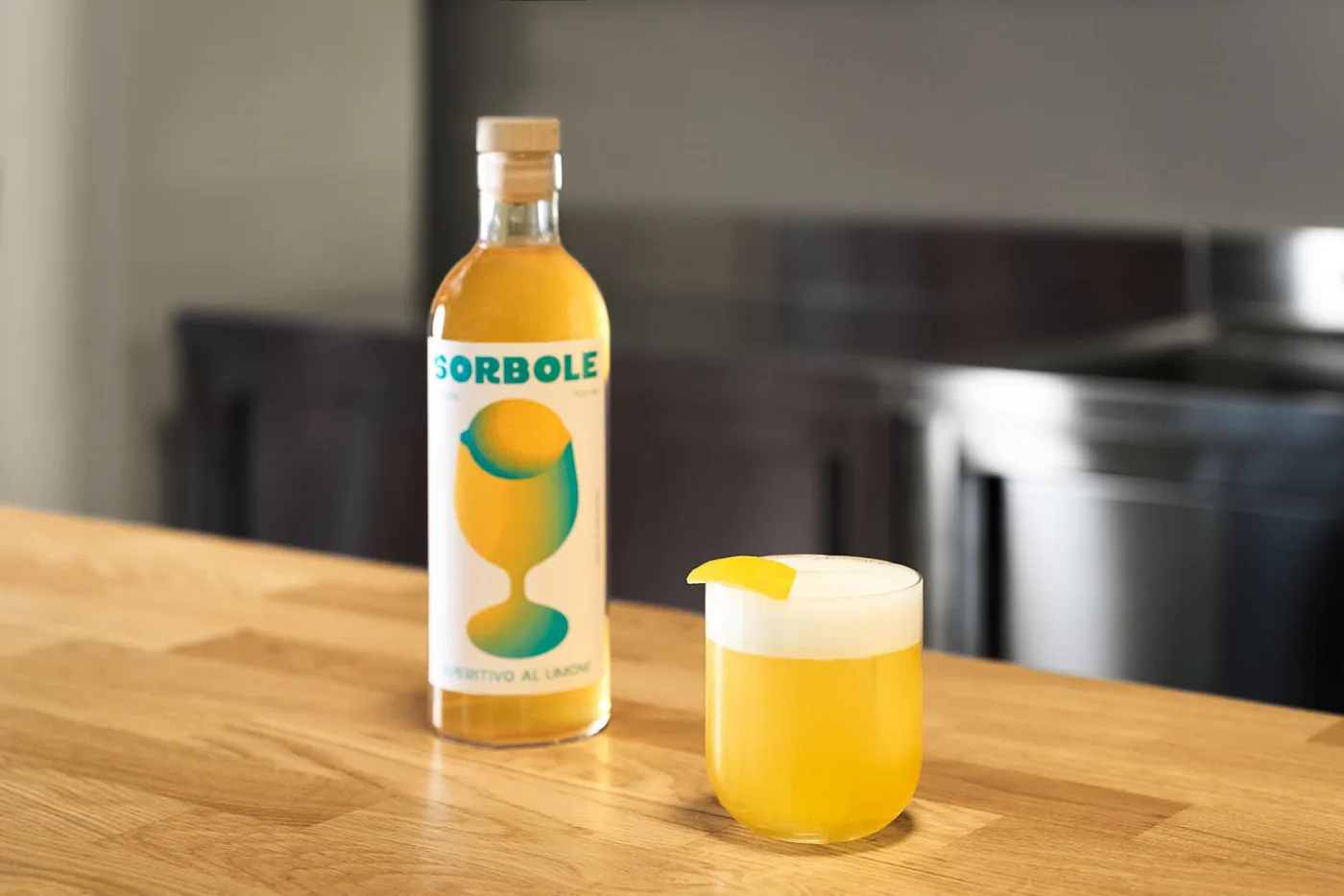 Sorbole Sour in tumbler basso con schiuma bianca e scorza di limone accanto alla bottiglia di Sorbole