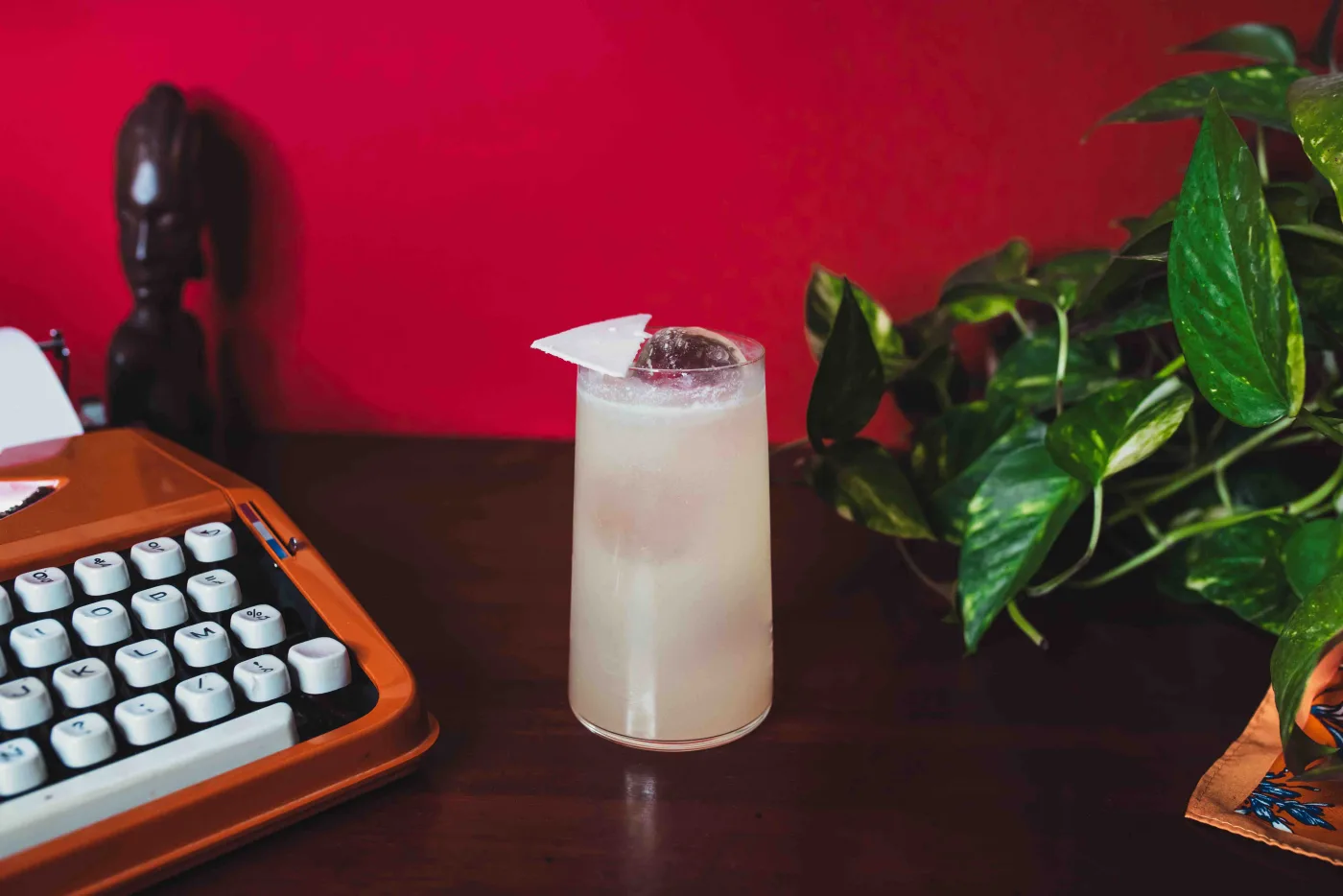 Thai Trip: long drink lattiginoso in tumbler alto con garnish di cocco, accanto a una macchina da scrivere vintage su fondo rosso