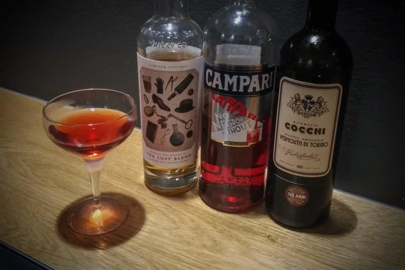 The Lost Boulevardier: coppetta con drink rosso accanto alle bottiglie di The Lost Blend, Campari e vermouth Cocchi