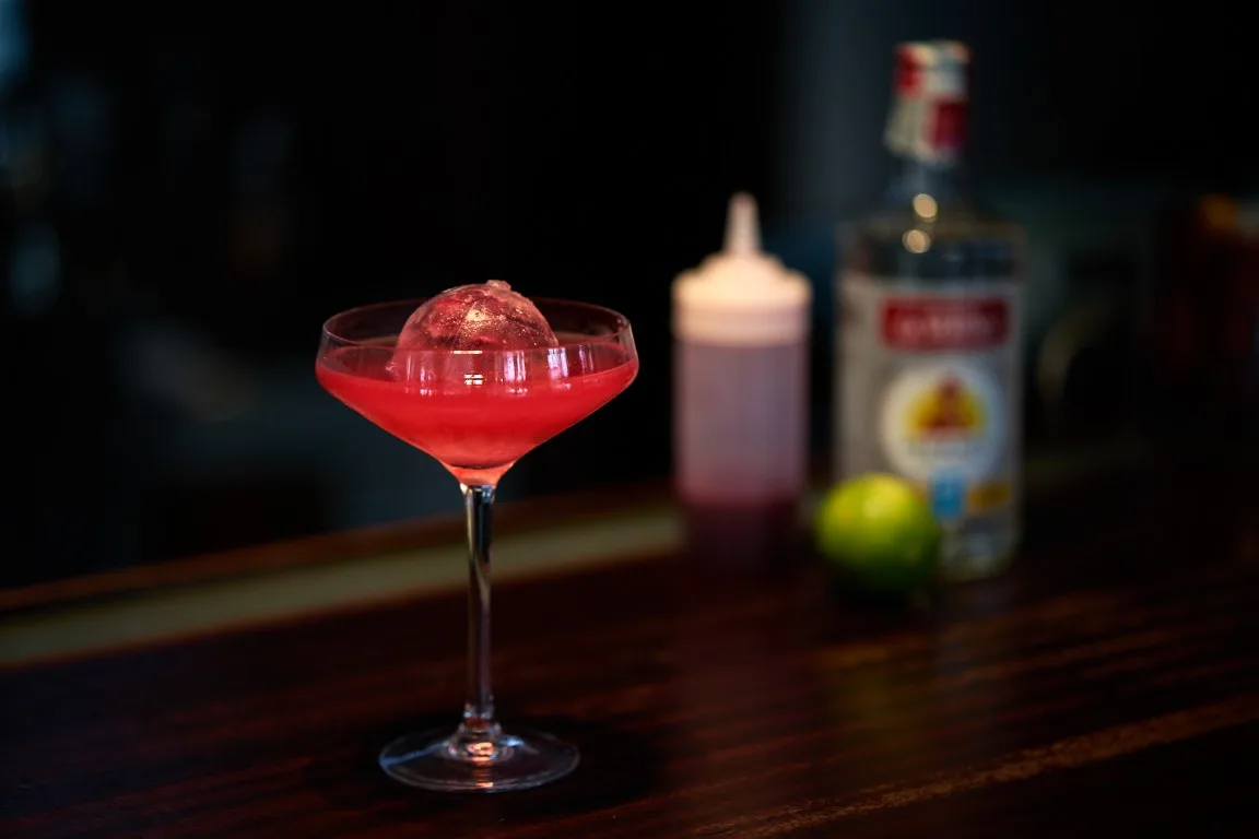 Violet Isle: coppetta con drink rosso acceso e sfera di ghiaccio ai petali di rosa, con squeezer di granatina e rum sullo sfondo
