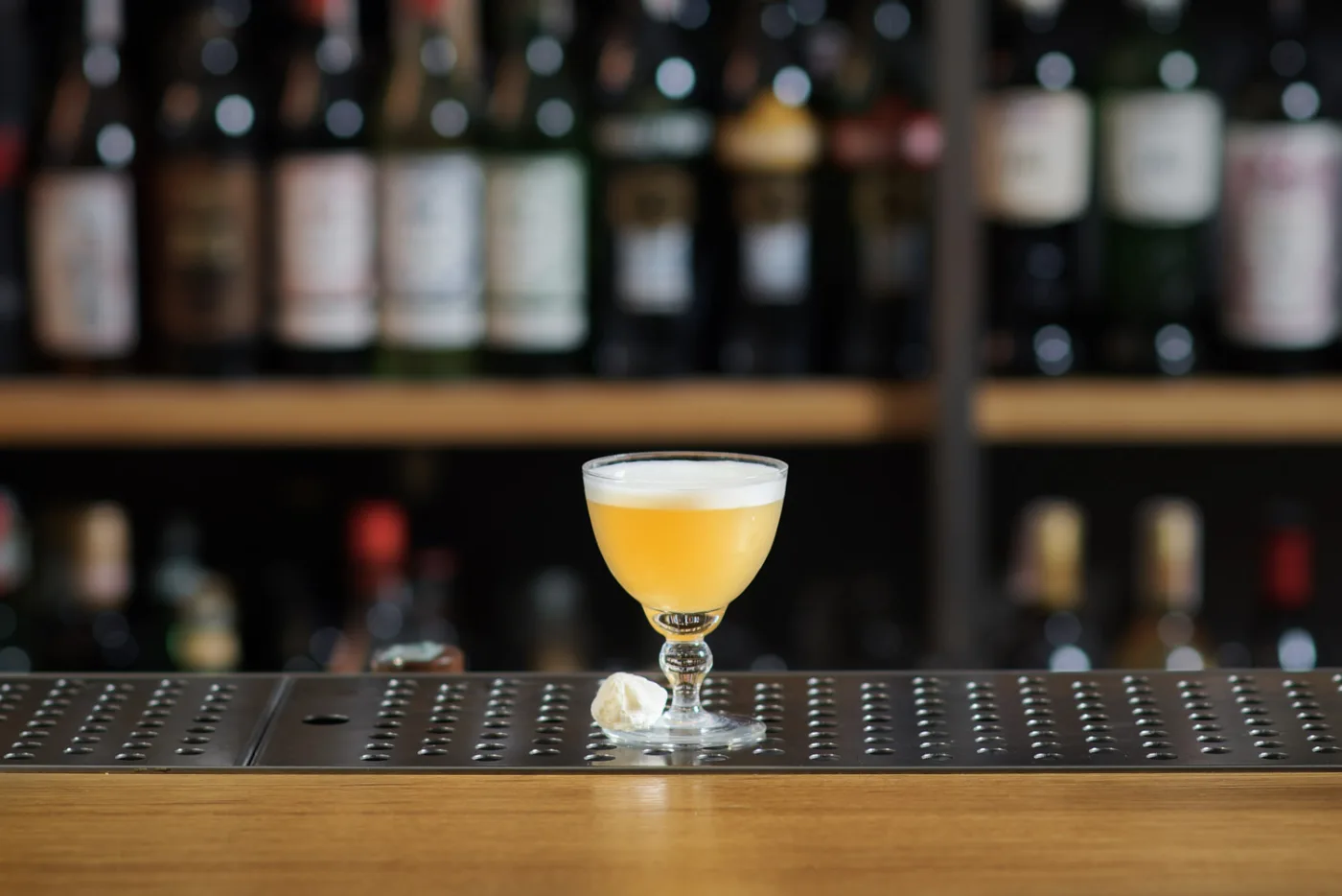 Il Whiskey Sour con la sua schiuma compatta servito in una coppetta vintage, davanti alla bottigliera del bar