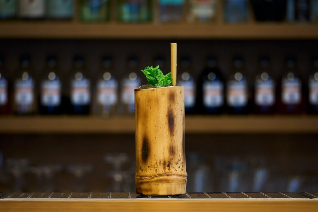 Su un banco bar, il cocktail Tiki Big Bamboo all'interno di un bicchiere inserito in una grossa canna di bamboo. Il cocktail è decorato con un rametto di menta