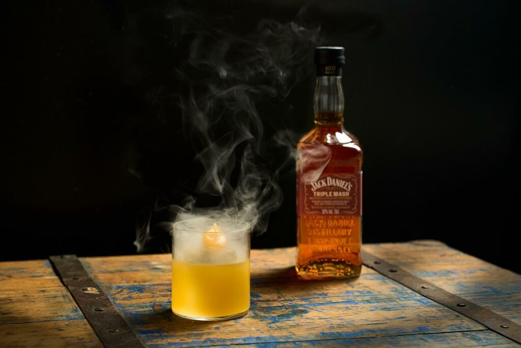 Su un baule di legno vintage il cocktail Out of Penicillin Dal bicchiere fuoriesce del fumo dato dall'affumicatura. Sullo sfondo una bottiglia di Jack Daniel's Triple Mash