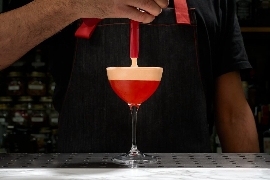 su un banco bar il cocktail Crème de la Crêpe servito in una coppetta da cocktail. si intravede il sifone che eroga la foam di crepe alla nutella in superficie al drink.