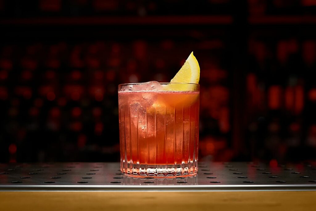 Su un banco bar un bicchiere tumbler basso contenente il cocktail Fellini decorato con uno spicchio di limone.
